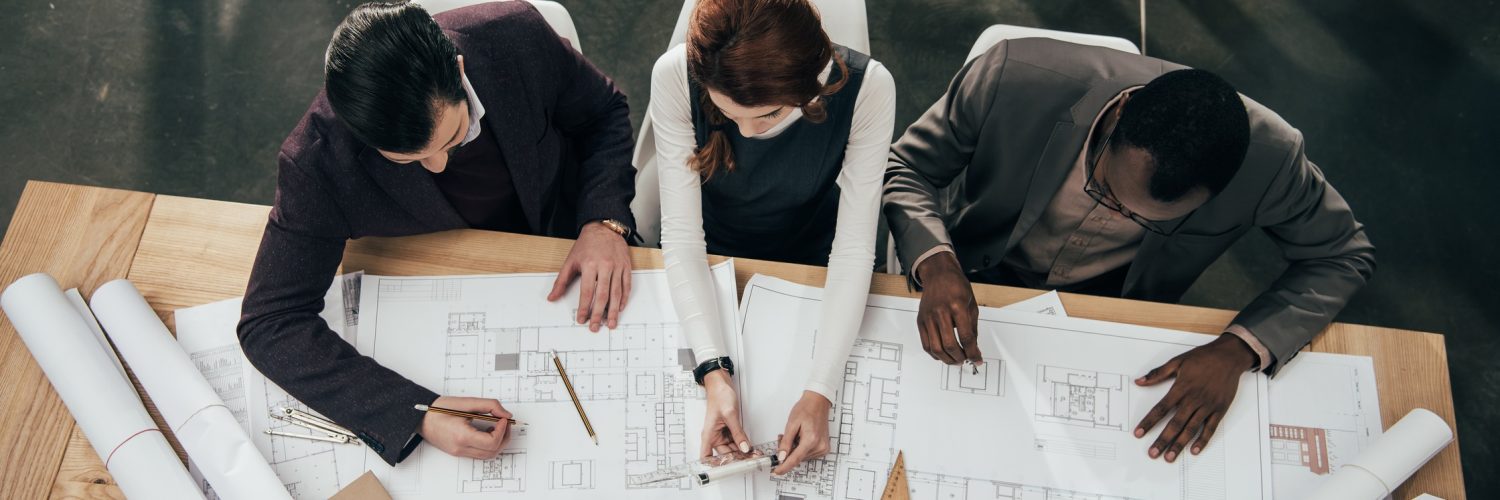 Three architects work together at a large table in an office and make notes on copies of blueprint plans.