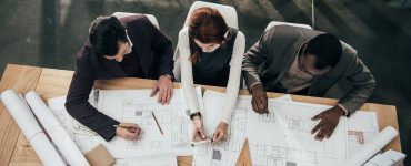 Three architects work together at a large table in an office and make notes on copies of blueprint plans.