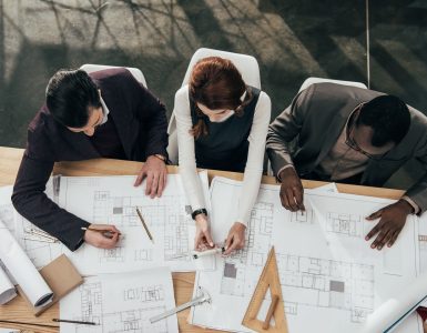 Three architects work together at a large table in an office and make notes on copies of blueprint plans.