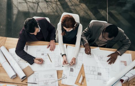 Three architects work together at a large table in an office and make notes on copies of blueprint plans.