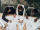 Three architects work together at a large table in an office and make notes on copies of blueprint plans.