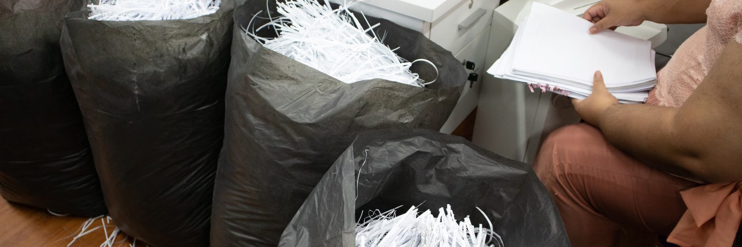 An office employee sorting and shredding confidential business paperwork. Some shreds are in large black trash bags.