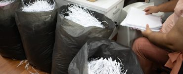 An office employee sorting and shredding confidential business paperwork. Some shreds are in large black trash bags.