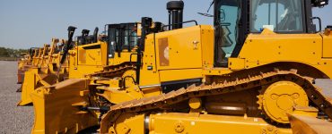 A row of yellow bulldozers parked on a construction site. The ground is flat with a layer of gravel.