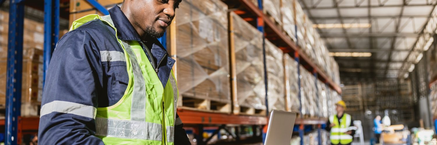 A man in a safety vest working on a laptop inside a warehouse. Behind him are shelves holding plastic-wrapped boxes.
