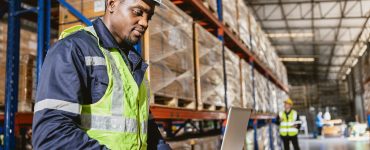 A man in a safety vest working on a laptop inside a warehouse. Behind him are shelves holding plastic-wrapped boxes.