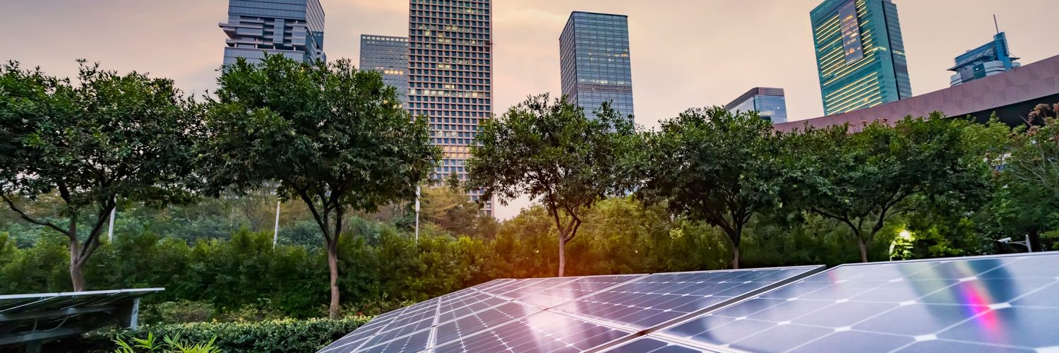 Solar panels on top of the roof of a building surrounded by various trees and multiple tall skyscrapers in the background.