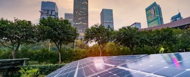 Solar panels on top of the roof of a building surrounded by various trees and multiple tall skyscrapers in the background.