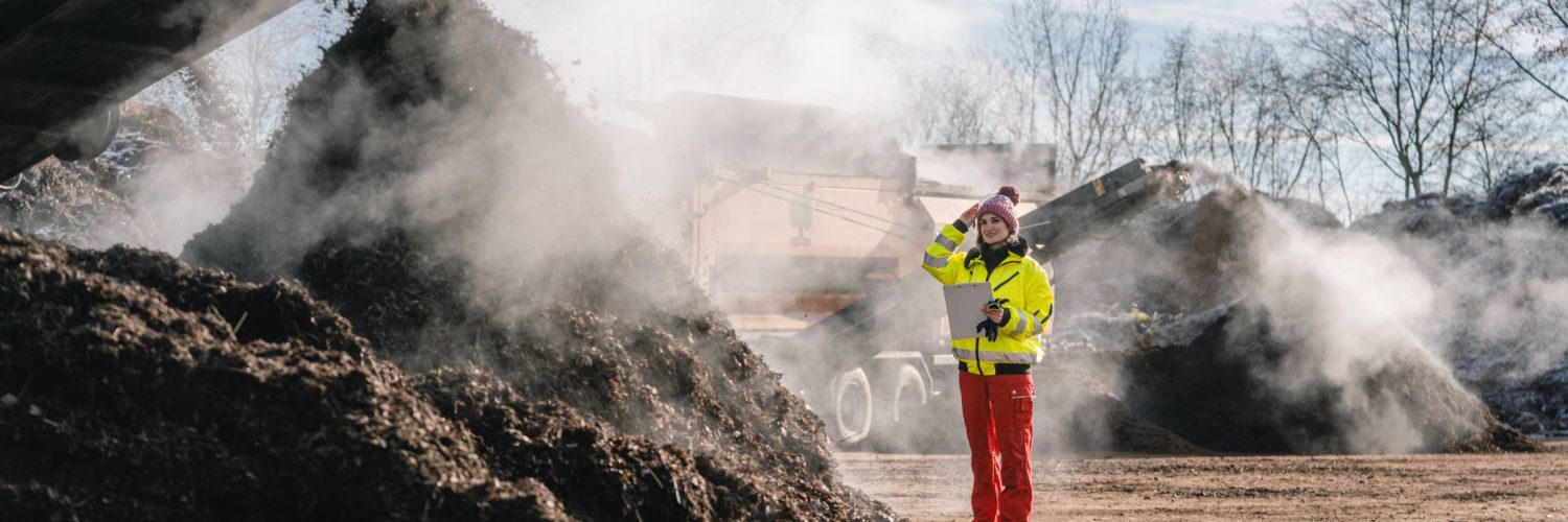 A female business professional standing near a pile of compost that's being added to by an excavator attachment.