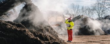 A female business professional standing near a pile of compost that's being added to by an excavator attachment.