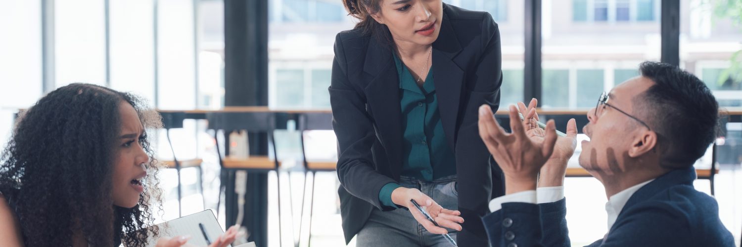 A seated woman holds a notebook, a standing woman leans on the table, and a man raises his hands while seated.