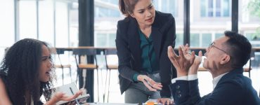 A seated woman holds a notebook, a standing woman leans on the table, and a man raises his hands while seated.
