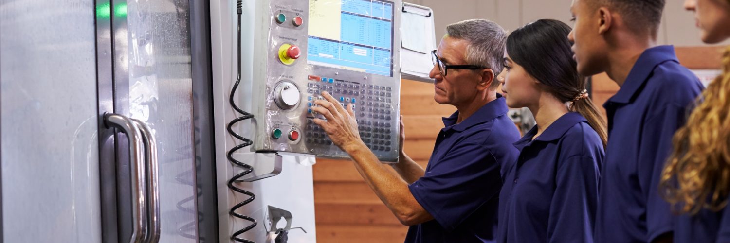 A gray-haired man in navy presses buttons on a CNC machine panel while three others in navy stand nearby watching.