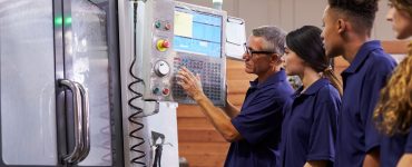 A gray-haired man in navy presses buttons on a CNC machine panel while three others in navy stand nearby watching.