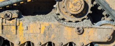 A close-up shows a bulldozer’s undercarriage with dried mud on the tracks and gears, lit by natural light.