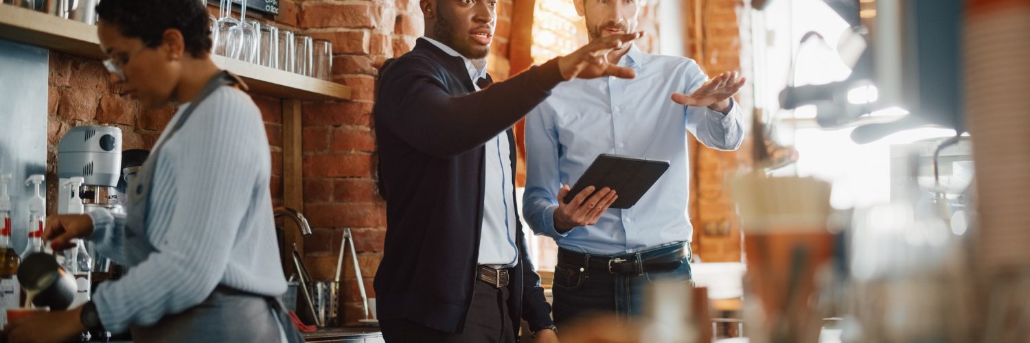 Two coffee shop male owners are meeting behind the counter, using a tablet in a cozy loft-style cafe.