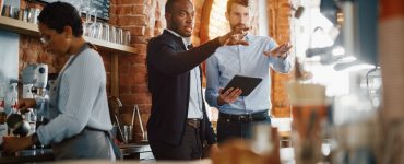 Two coffee shop male owners are meeting behind the counter, using a tablet in a cozy loft-style cafe.