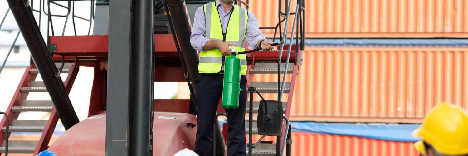 A man standing on a crane car while holding a fire extinguisher. There are employees watching him from the ground.