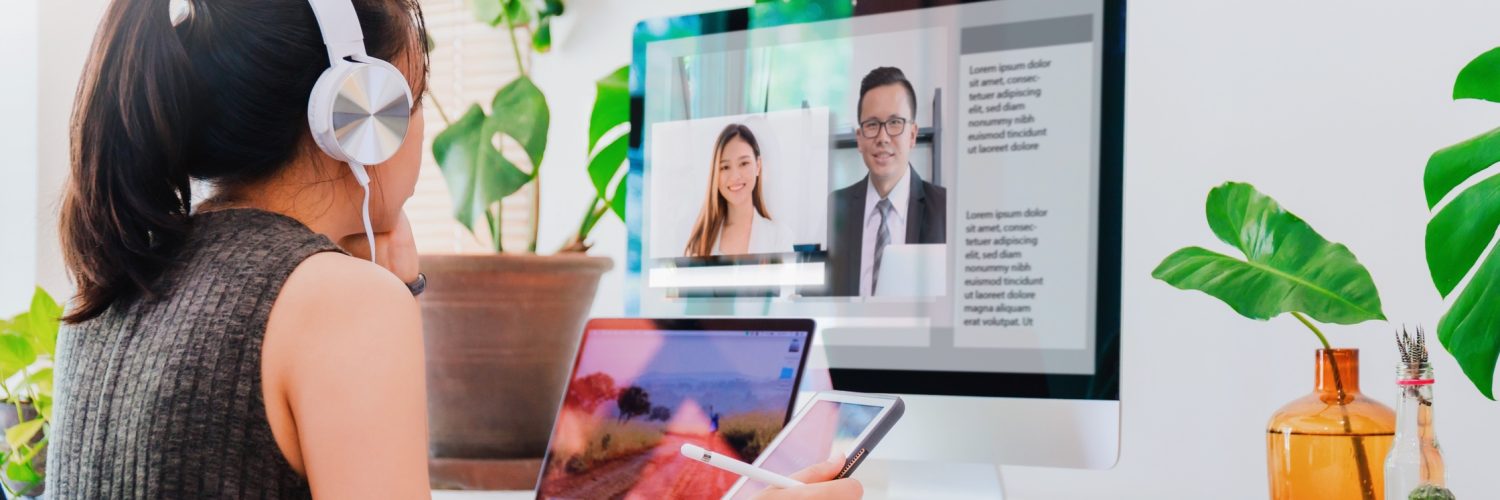 A woman sits at a desk with a laptop and monitor at eye level. She's on a video call, wearing headphones and holding a tablet.
