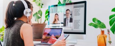 A woman sits at a desk with a laptop and monitor at eye level. She's on a video call, wearing headphones and holding a tablet.