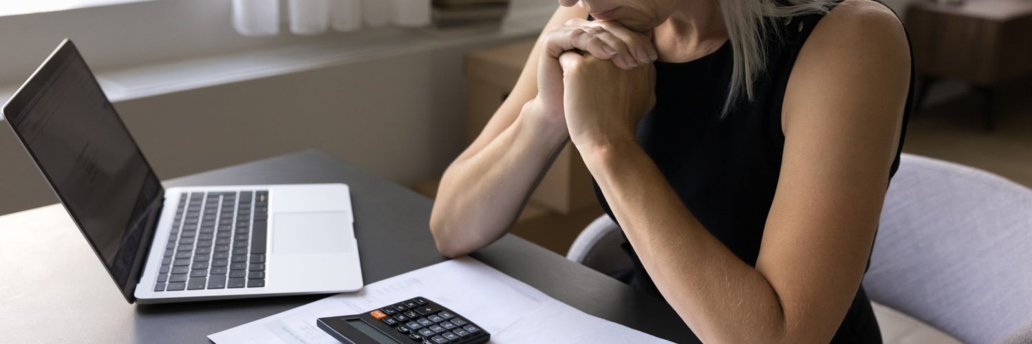 A woman rests her elbows on a gray desk while reviewing printed data with a laptop and a black calculator within reach.