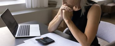 A woman rests her elbows on a gray desk while reviewing printed data with a laptop and a black calculator within reach.