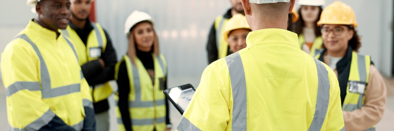 A group of workers paying attention to someone holding a tablet. They are all wearing safety vests and hard hats.