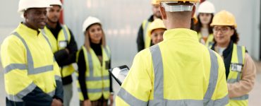 A group of workers paying attention to someone holding a tablet. They are all wearing safety vests and hard hats.