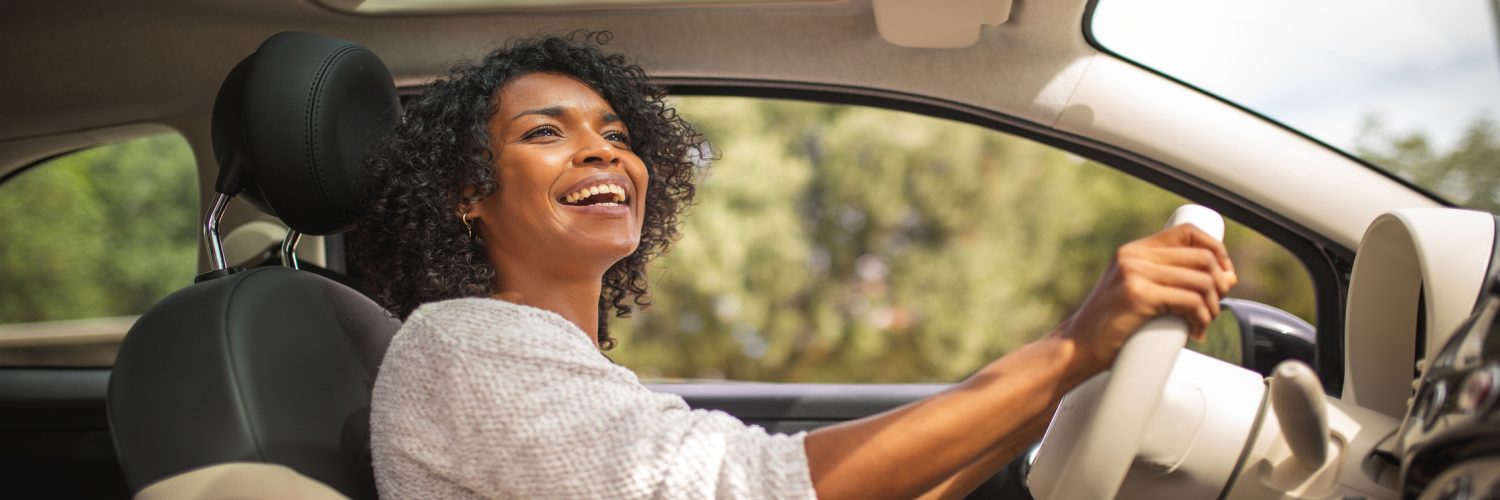 A smiling young Black woman is driving her car with her sunroof open. She's looking up at her rear-view mirror.
