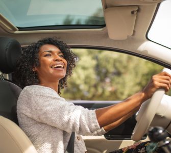 A smiling young Black woman is driving her car with her sunroof open. She's looking up at her rear-view mirror.