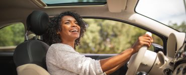 A smiling young Black woman is driving her car with her sunroof open. She's looking up at her rear-view mirror.