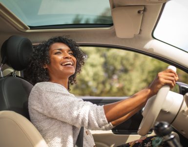 A smiling young Black woman is driving her car with her sunroof open. She's looking up at her rear-view mirror.