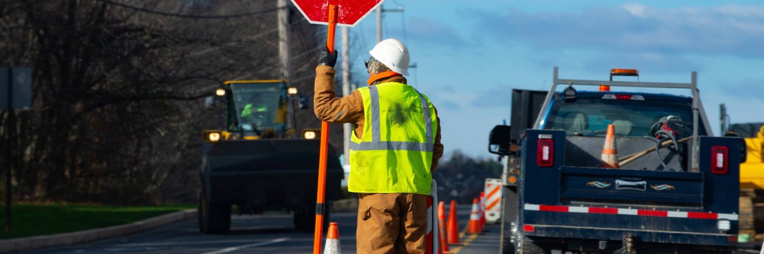 A construction worker in a safety vest stands on the road next to orange traffic cones holding a stop sign.