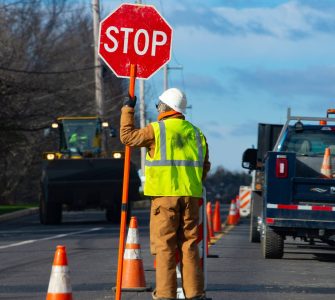 A construction worker in a safety vest stands on the road next to orange traffic cones holding a stop sign.