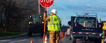 A construction worker in a safety vest stands on the road next to orange traffic cones holding a stop sign.