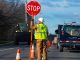 A construction worker in a safety vest stands on the road next to orange traffic cones holding a stop sign.