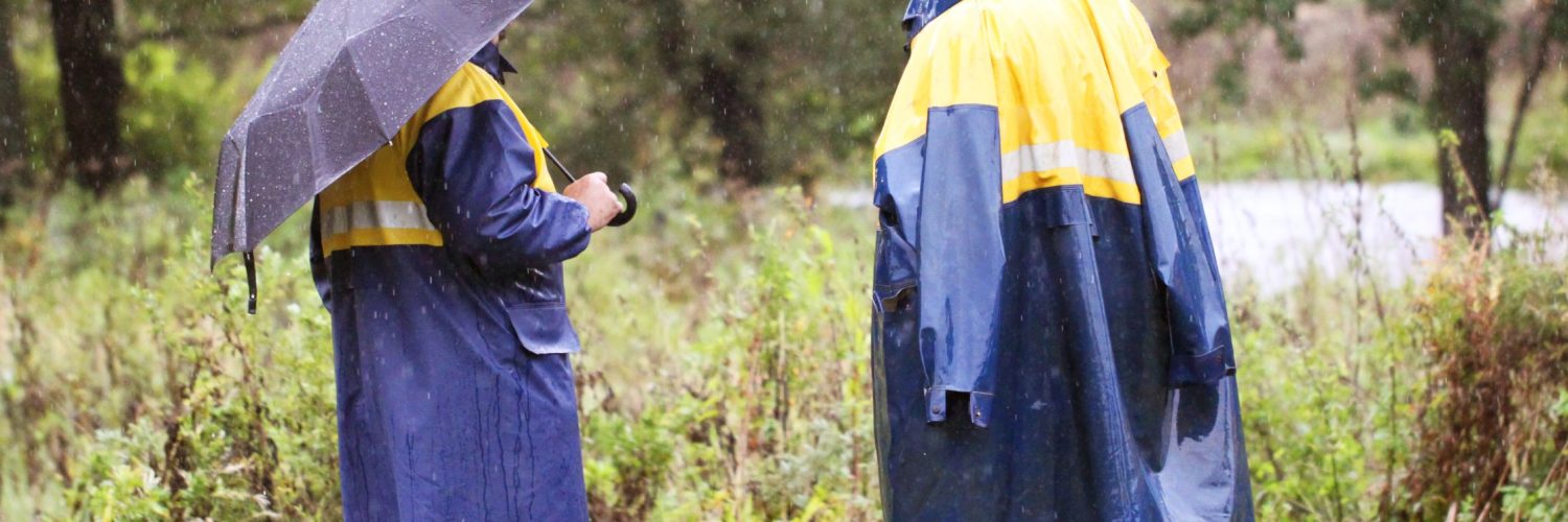 Two workers stand in the rain wearing high-visibility rain coats, one with a hood, the other with an umbrella.