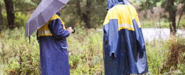 Two workers stand in the rain wearing high-visibility rain coats, one with a hood, the other with an umbrella.