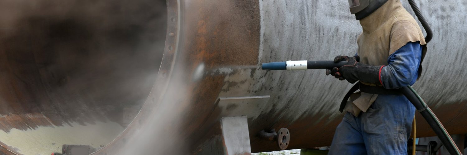 An industrial worker in a protective suit with a metal helmet holds a sandblasting gun toward a metal cylinder outside.