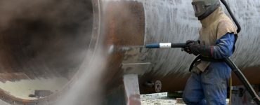 An industrial worker in a protective suit with a metal helmet holds a sandblasting gun toward a metal cylinder outside.