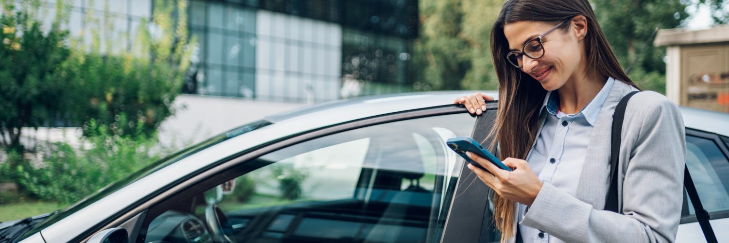 A person wearing glasses and a business suit smiling while standing next to a car and looking at their phone.