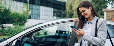A person wearing glasses and a business suit smiling while standing next to a car and looking at their phone.
