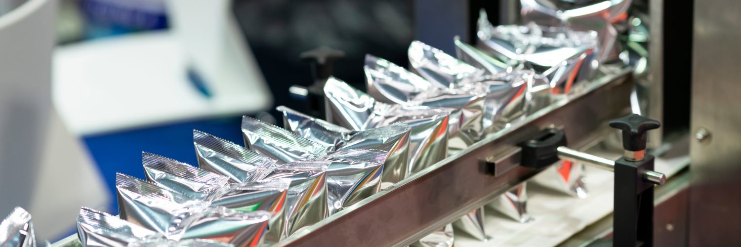 A machine holds several shiny, silver plastic pouches lined up in a row and secured by a metal arm bar.