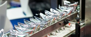 A machine holds several shiny, silver plastic pouches lined up in a row and secured by a metal arm bar.
