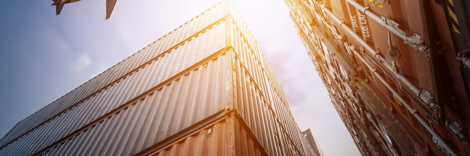 A low-angle shot of towering red and blue cargo containers. There is an airplane flying overhead in the sky.