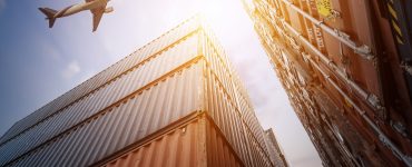 A low-angle shot of towering red and blue cargo containers. There is an airplane flying overhead in the sky.