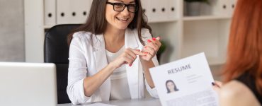 A manager grins at a job candidate who sits in front of her. The candidate holds a copy of their resume.