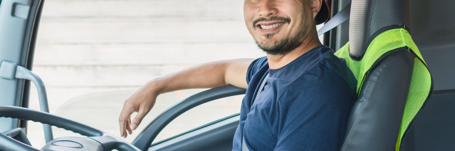A man wearing a navy shirt and hat smiles inside a truck with a gray interior. There's also a yellow vest over his seat.