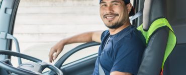 A man wearing a navy shirt and hat smiles inside a truck with a gray interior. There's also a yellow vest over his seat.