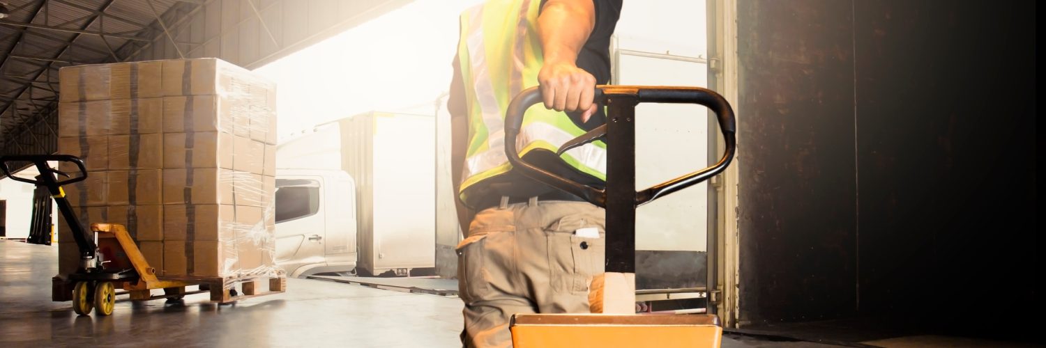 A man pulling a pallet jack toward a docking station at a warehouse. There are boxes wrapped in plastic on the equipment.